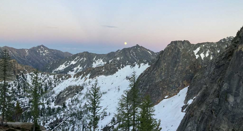 A full moon appears above snowy mountains in a sky illuminated in soft blues and purples. 